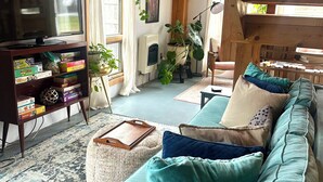A cozy living room with a wooden ceiling, a television on a stand, a bookshelf, and a teal sofa with cushions.