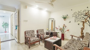 A spacious living room with a high ceiling fan, two patterned sofas, a coffee table, and a side table. There is a potted plant and a decorative tree mural on the wall. - Octave Church Inn (Bengaluru)