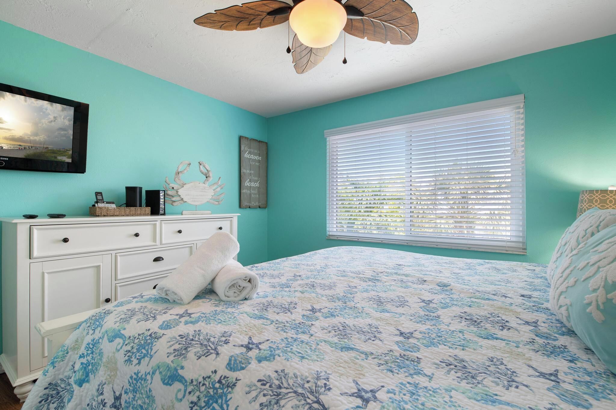 Un dormitorio con cama, cómoda, ventilador de techo, ventana con persianas e imagen enmarcada en la pared.