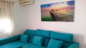 A teal sofa with multiple cushions in a room with a painting of a sunset over water on the wall. - Immaculate 2-bed Villa With Pool in Mazarrón (Mazarrón)