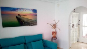 A teal sofa in a room with a sunset painting, a white door, and a small white shelf with a vase. - Immaculate 2-bed Villa With Pool in Mazarrón (Mazarrón)