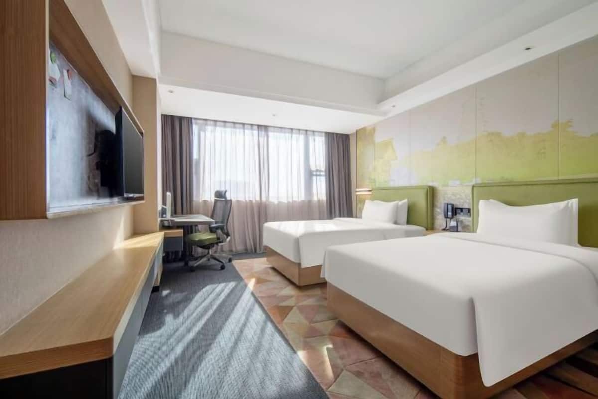 A hotel room with two beds, a desk, a chair, a TV, and a large window with curtains.