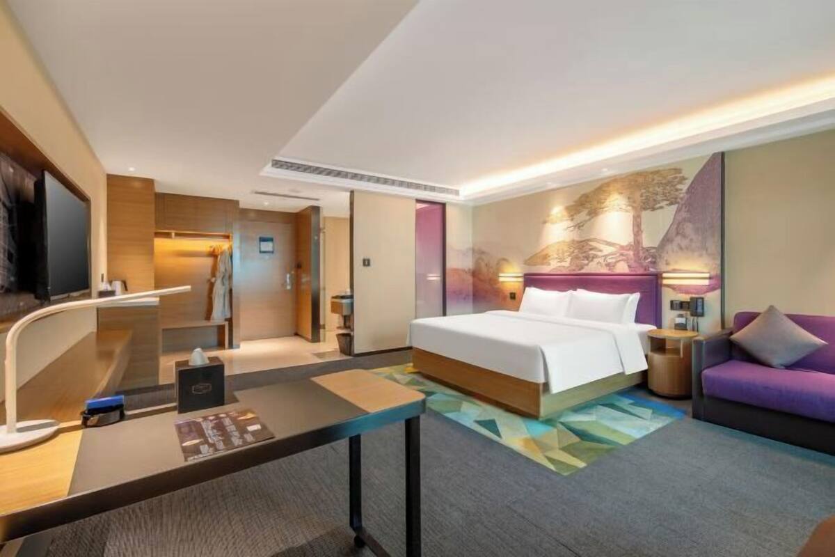 A hotel room with a bed, a desk, a sofa, a TV, and a wall with a painting.