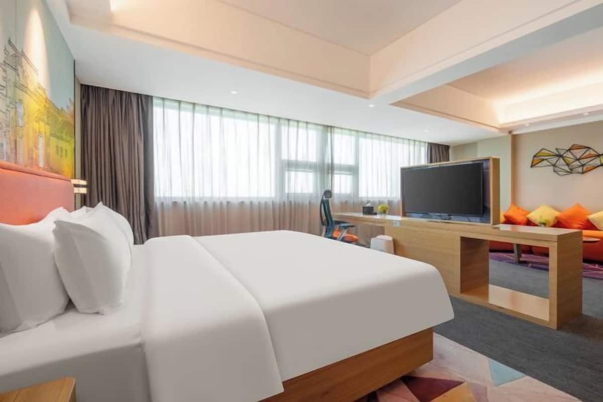 A hotel room with a bed, a desk with a TV, a painting on the wall, and a window with curtains.