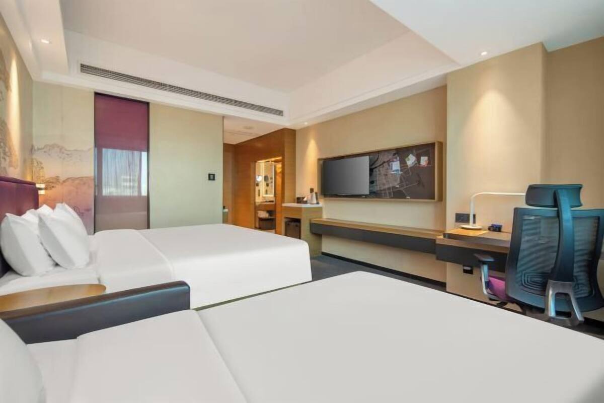 A hotel room with two beds, a desk, a chair, a TV, and a bathroom visible through an open door.