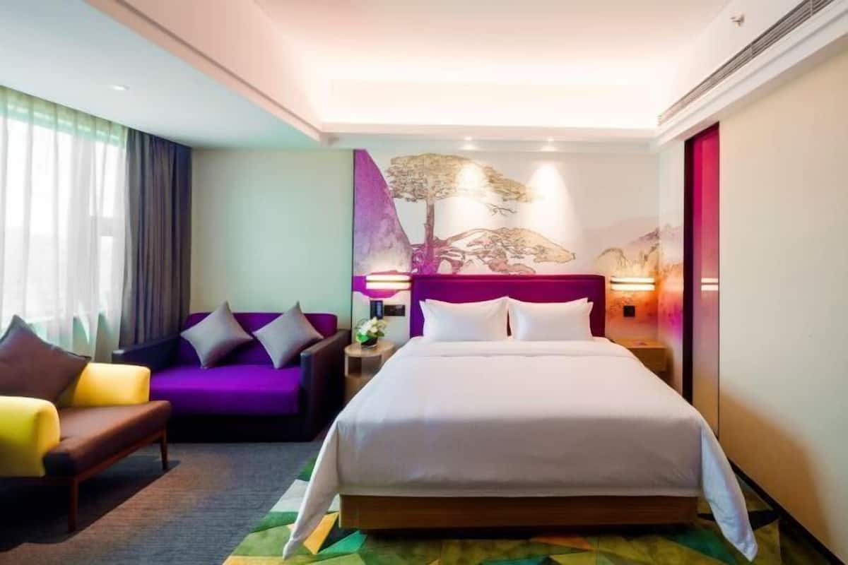 A hotel room with a large bed, a purple sofa, a yellow armchair, and a wall-mounted artwork of a tree.