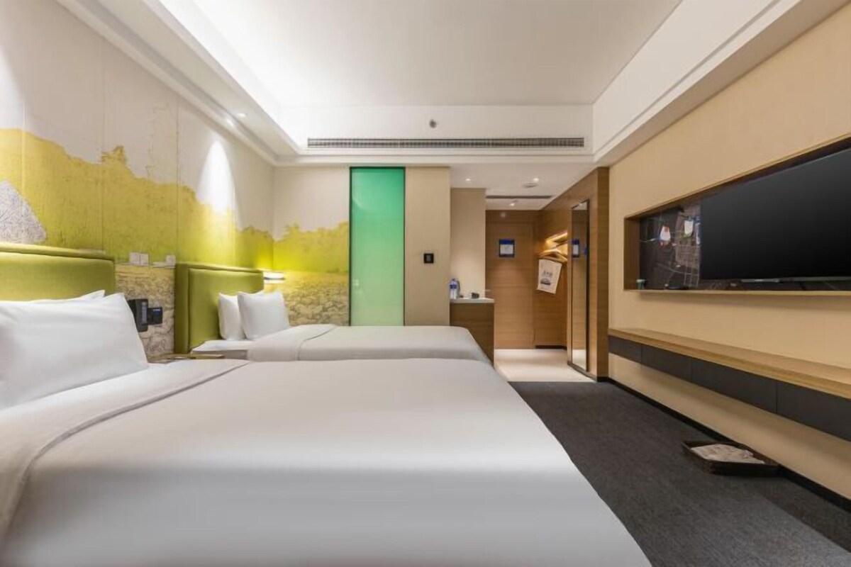 Habitación de hotel con dos camas, un televisor de pantalla plana grande montado en la pared, una puerta verde y un escritorio pequeño con una silla.