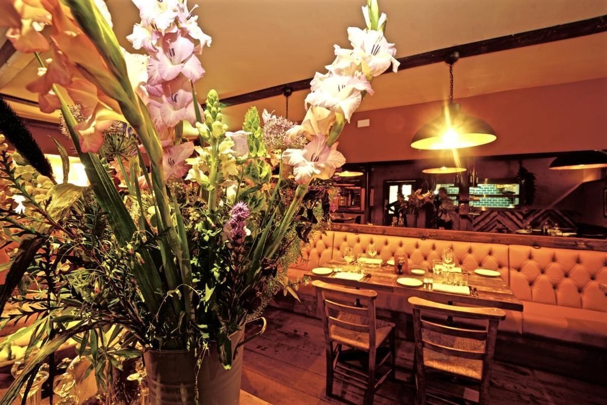 A restaurant with a wooden table and chairs, a cushioned bench, and a large vase of flowers.