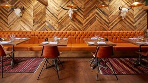 A restaurant with a herringbone wood wall, a central orange cushioned bench, and wooden tables with chairs. - The Bull Hotel (Fairford)