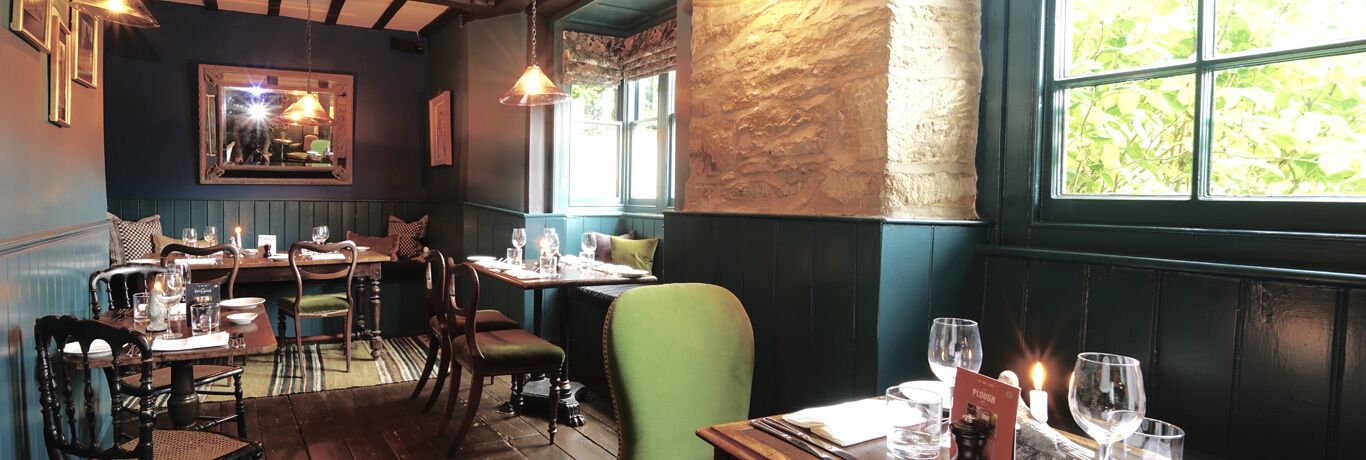 Een restaurantinterieur met houten meubels, een groen gestoffeerde bank en een groot raam met uitzicht op groen.