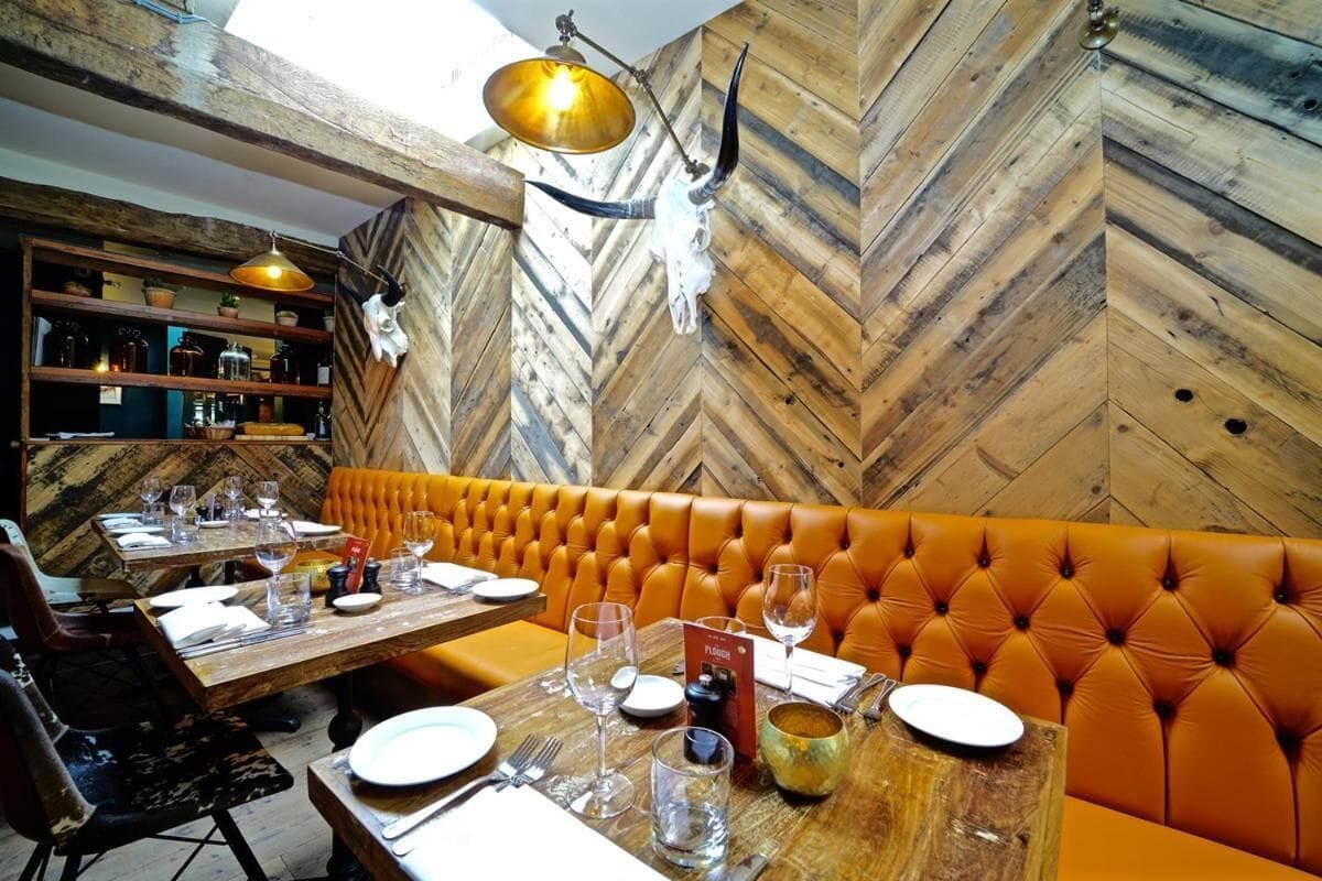 A restaurant interior with wooden herringbone walls, a tufted orange booth, and a wooden table set for dining.