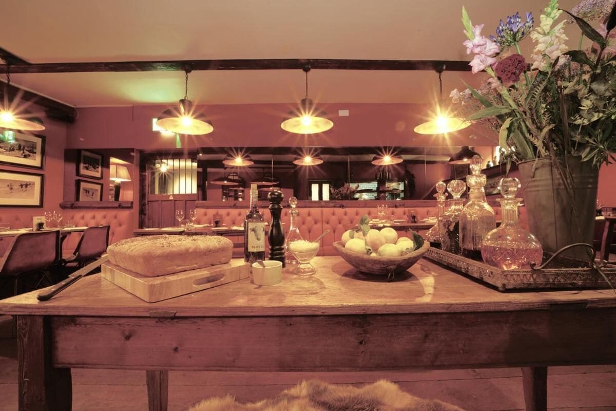 A bar area with wooden tables, hanging pendant lights, and a variety of bottles and glasses.