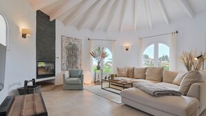 A spacious living room with a white sofa, a coffee table, and a fireplace. There is a large window with curtains and a view of the outdoors.