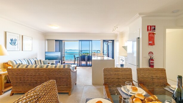 A modern living room with a large TV, wicker furniture, and a view of the sea. - Florentine, 14/11 Columbia Cl - – AIRCON, WIFI, UNSURPASSED WATER VIEWS (Nelson Bay)