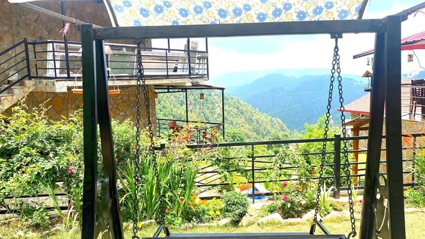 Une vue depuis un balcon doté d’une structure métallique, donnant sur une vallée verdoyante et des montagnes lointaines.