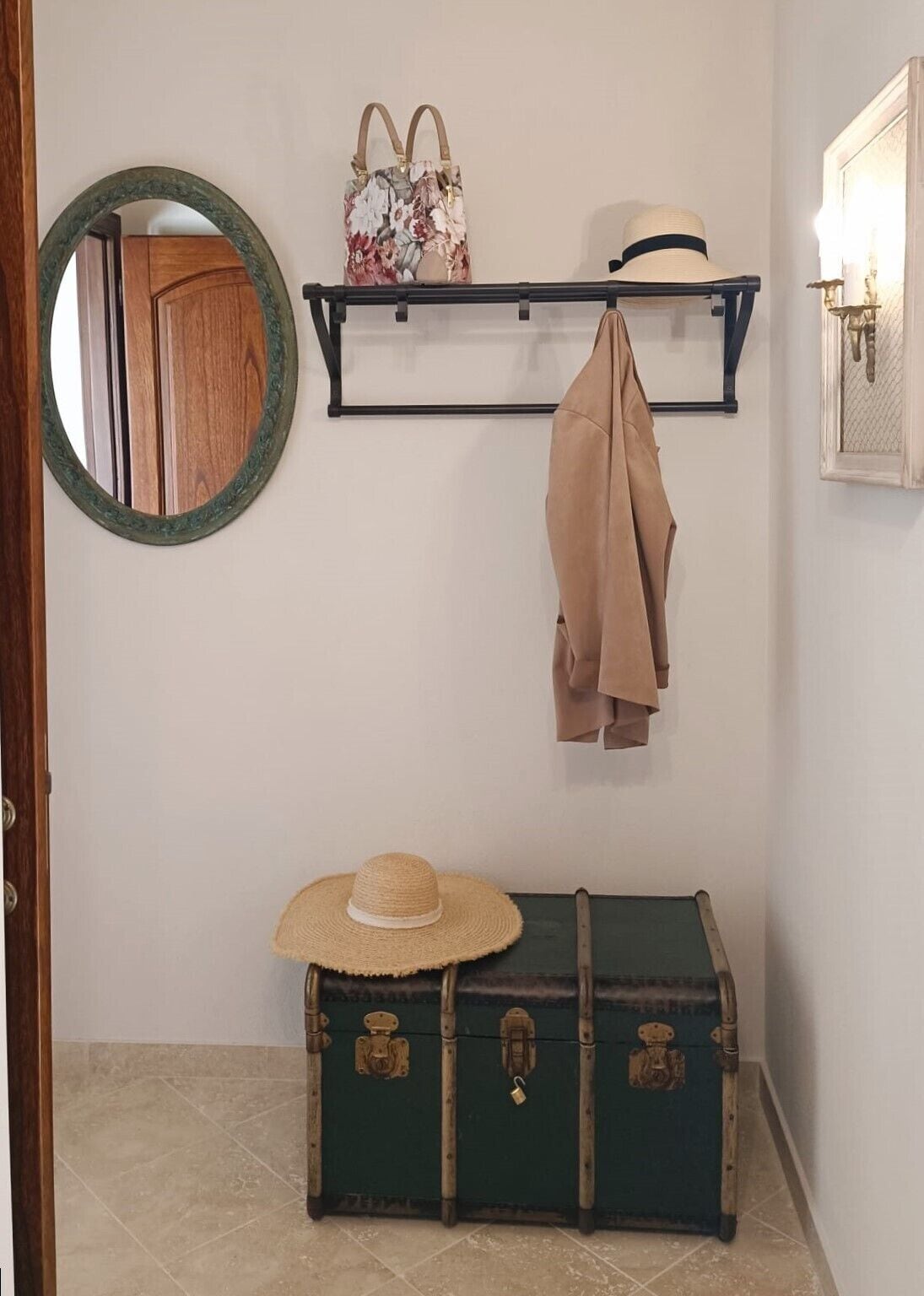 Une pièce avec une porte en bois, un miroir, une malle verte, un chapeau, une étagère avec un sac à main et un autre chapeau accroché au mur.