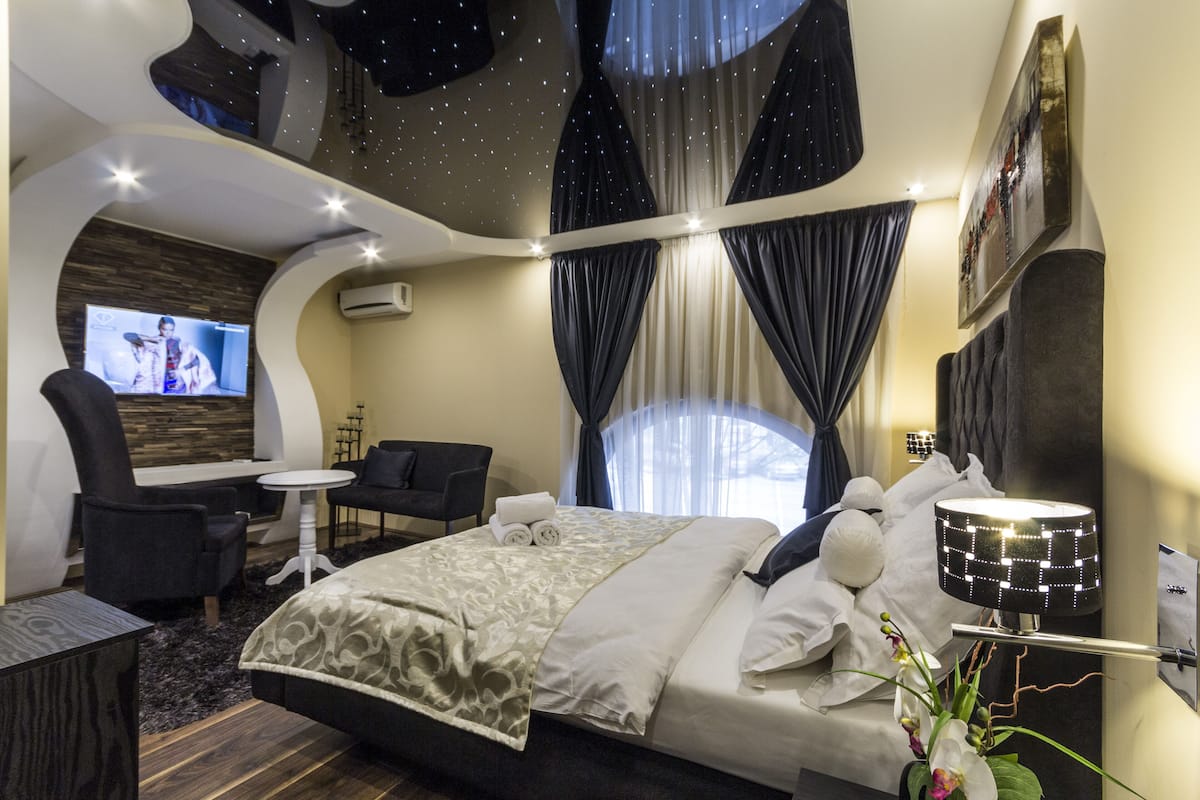 A modern hotel room with a large bed, a TV, a seating area with chairs, and a decorative ceiling.