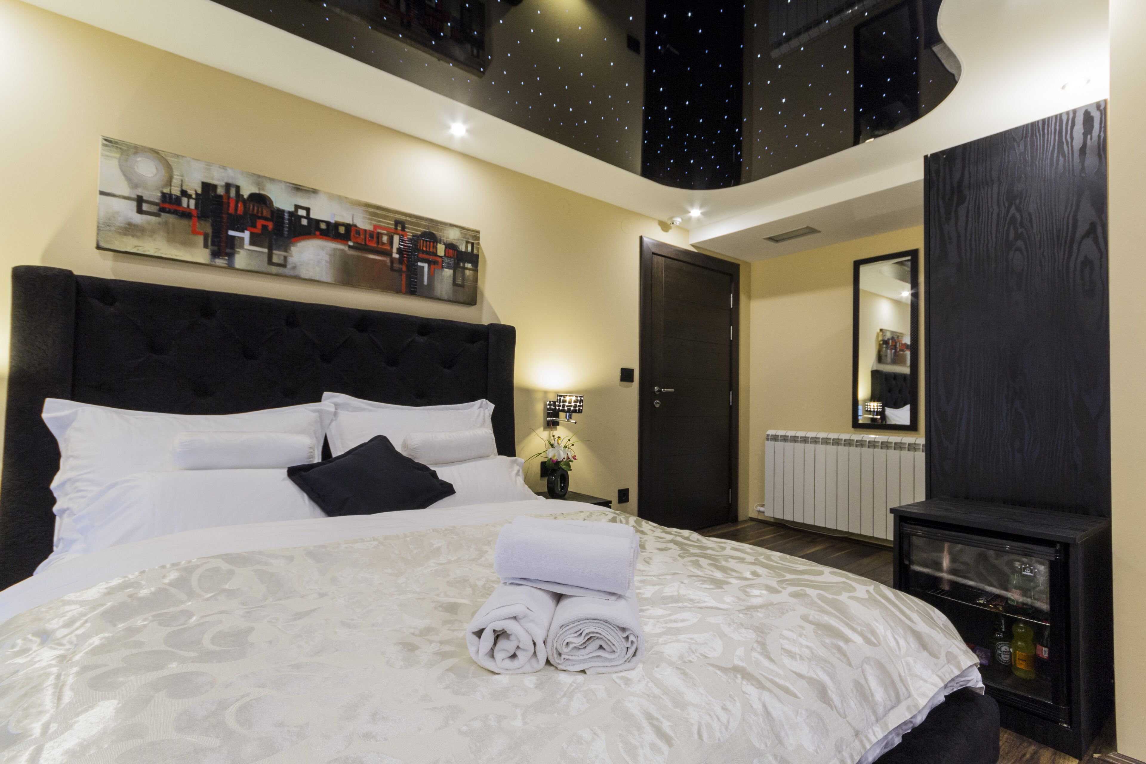 A hotel room with a large bed, a cityscape painting, a starry ceiling, a dark wooden cabinet, and a mirror on the wall.