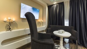 A modern hotel room with a flat-screen TV mounted on the wall, a cozy seating area with two chairs and a small round table, and a large window with black curtains. - Boutique Hotel Madison  (Belgrade)