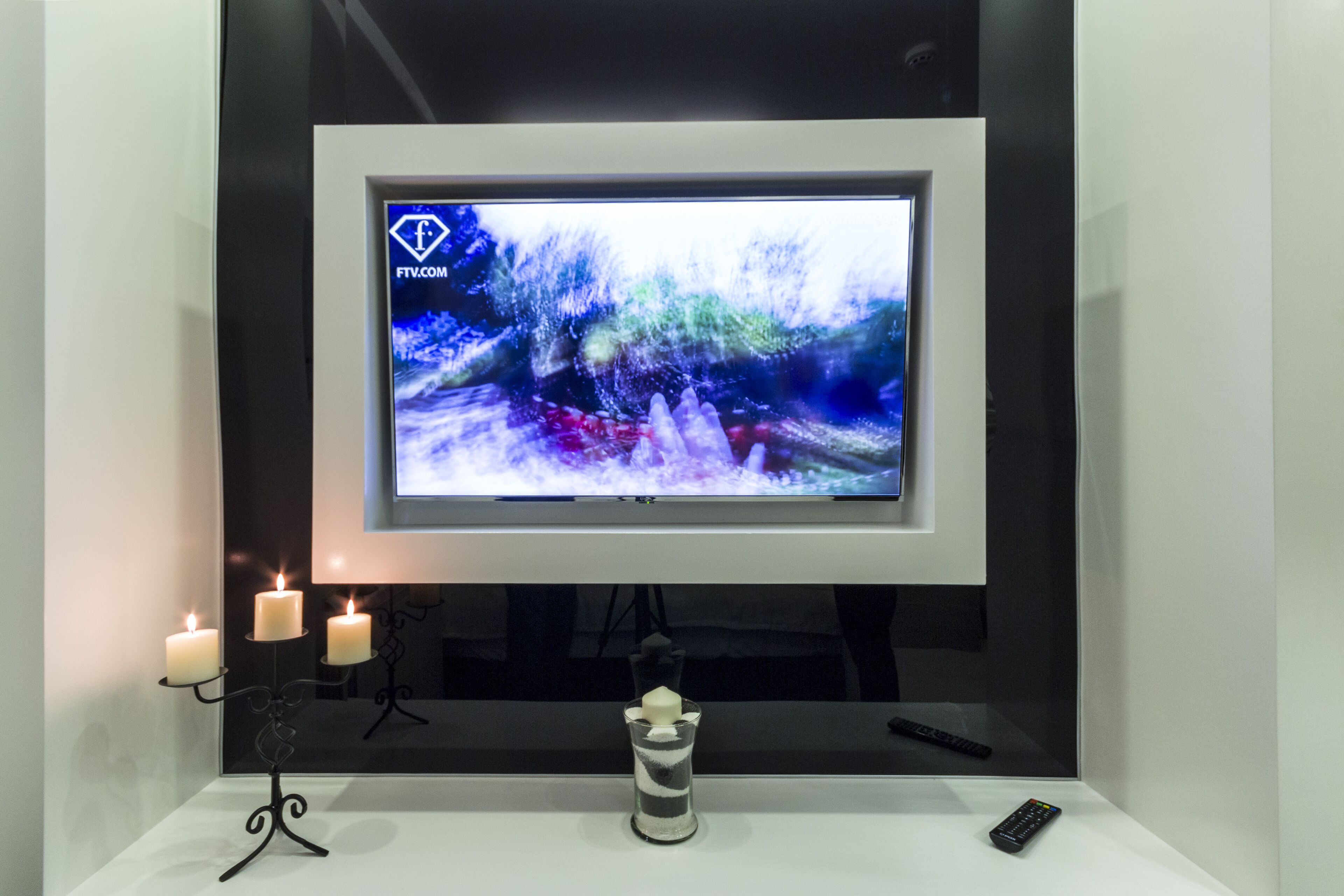 Ein modernes Wohnzimmer mit einem an der Wand befestigten Flachbildfernseher, einem kleinen Tisch mit Kerzen und einer Fernbedienung sowie einer dekorativen Glasvase.