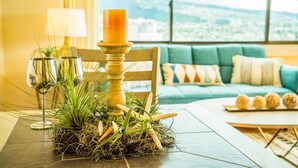A table with a candle, glasses, and a plant arrangement in a room with a view through a large window.