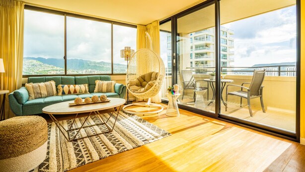 A modern living room with a sofa, a round coffee table, a wicker chair, and a balcony with a view of buildings and hills.