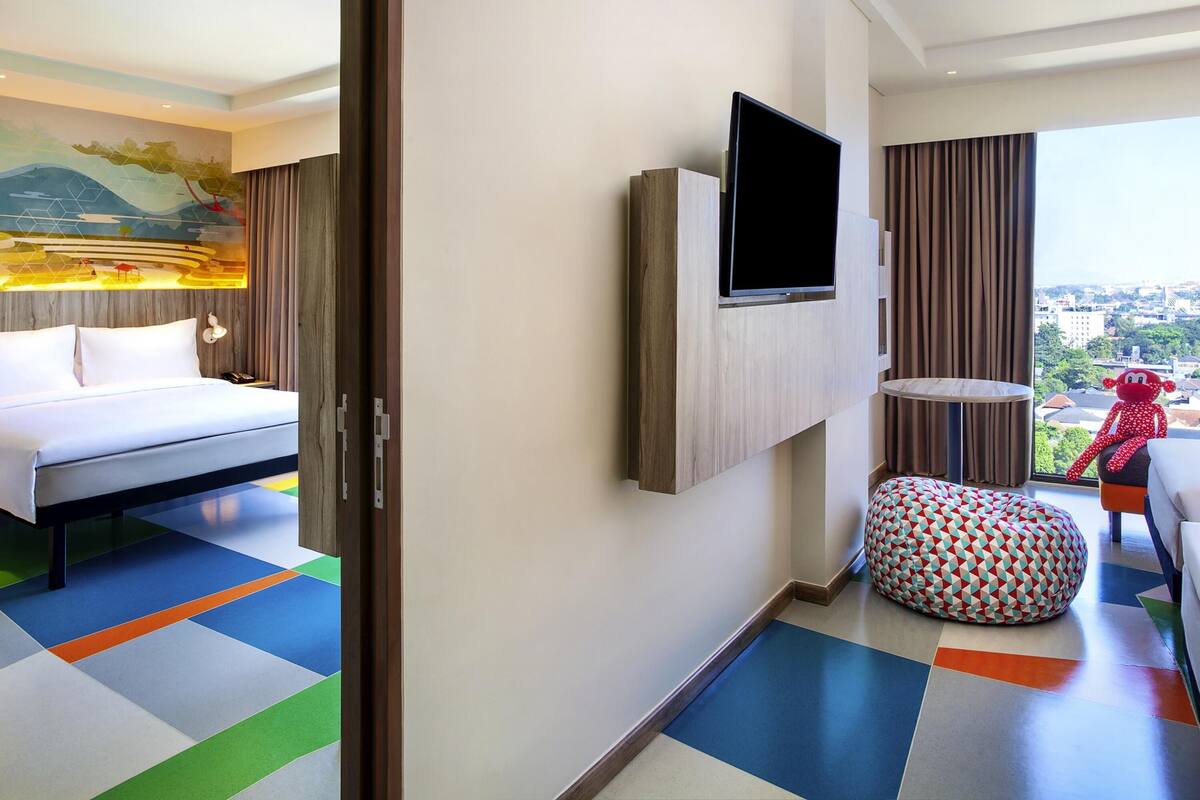 A hotel room with a bed, a mounted TV, a colorful floor, a window with a city view, and a red plush toy.