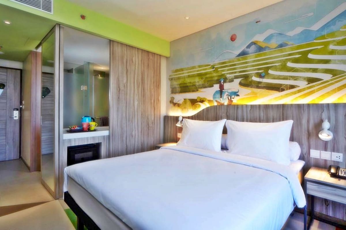A modern hotel room with a large bed, a bedside table with a lamp, a painting on the wall, and a sliding door leading to a bathroom.