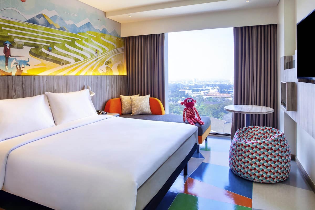 A modern hotel room with a large bed, a small round table, a bean bag chair, and a city view.