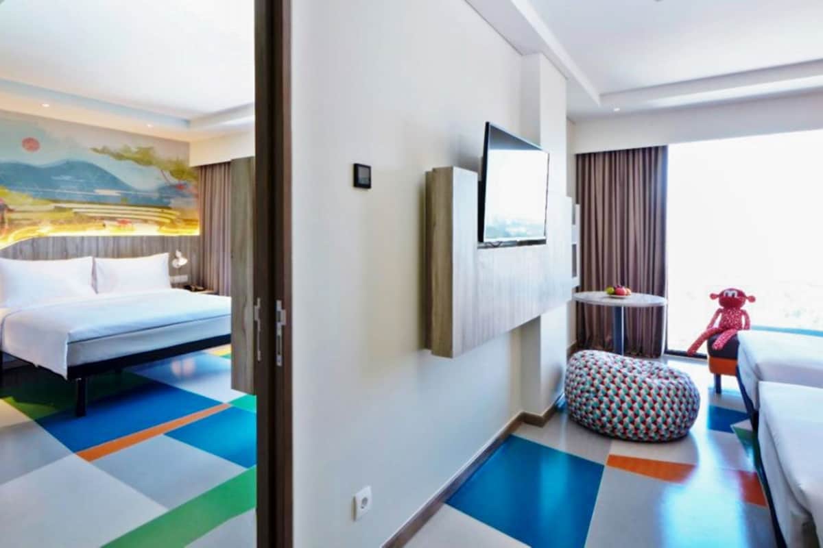 A hotel room with a bed, a TV, a small table with fruit, a colorful ottoman, and a mural on the wall.