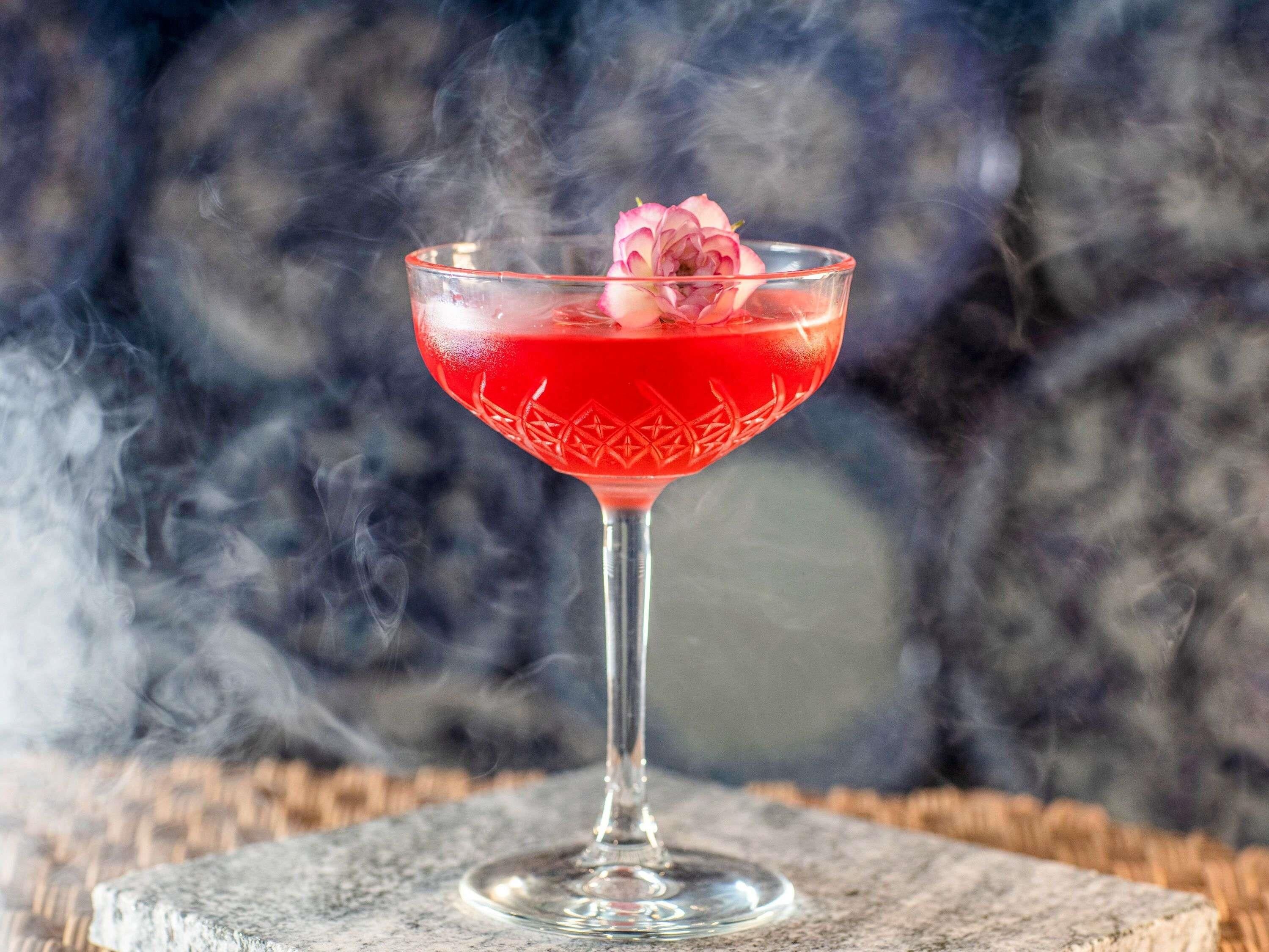 Un cocktail in un bicchiere con guarnizione di rosa, fumo che si alza, su sfondo sfocato.
