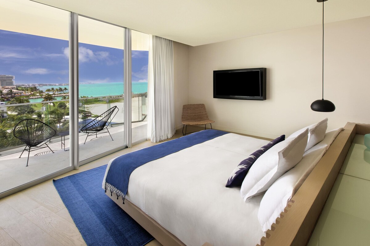 A hotel room with a bed, a TV, a chair, a balcony with a view of the ocean, and a large window.