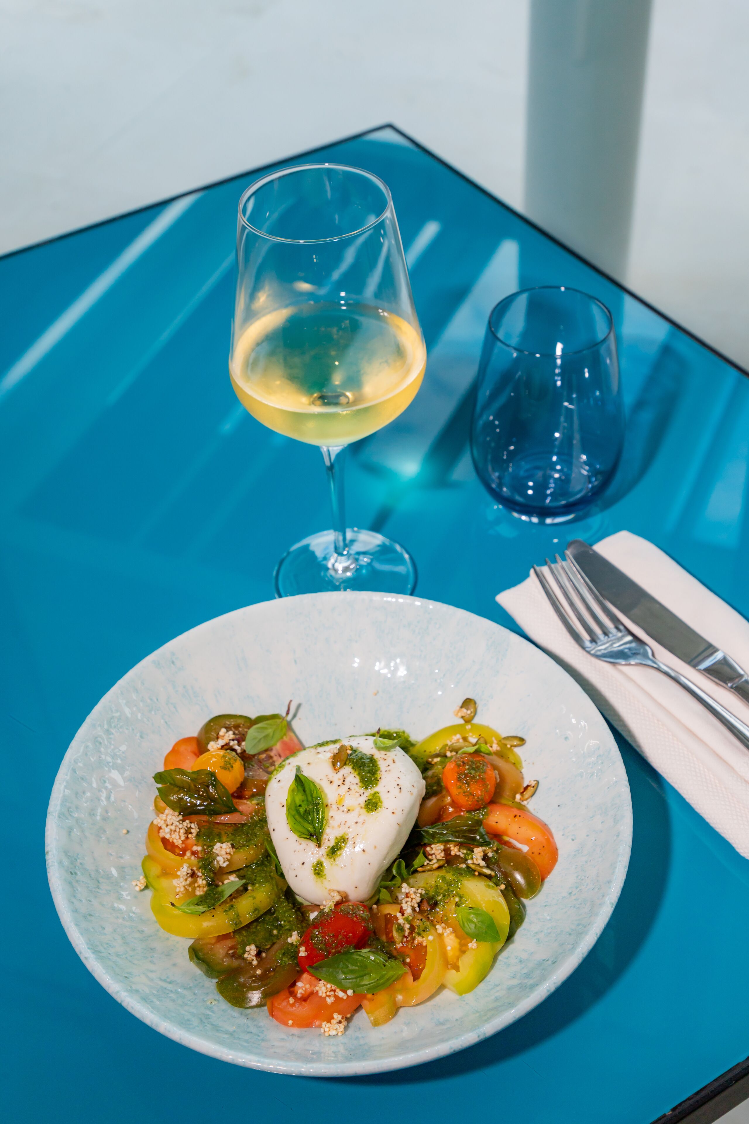 A plate of food with a white sauce and vegetables, a glass of white wine, and a napkin with cutlery on a blue table.