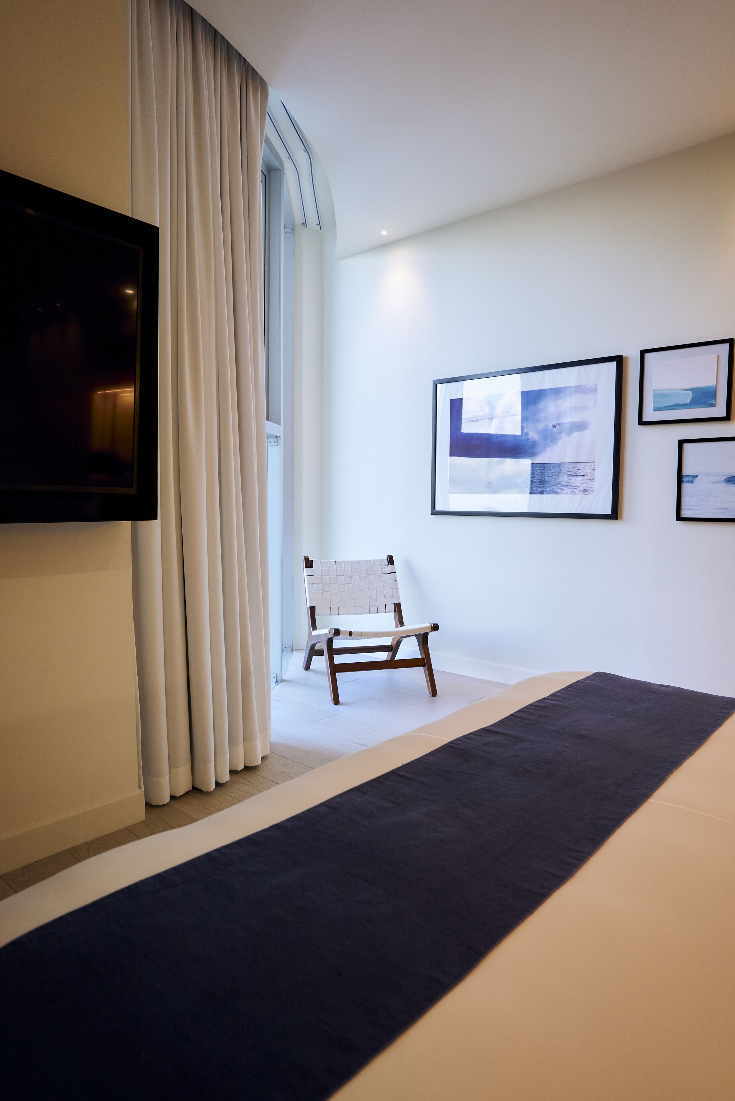A modern hotel room with a large window, a flat-screen TV, a bed with a dark blue bedspread, a white chair, and three framed pictures on the wall.