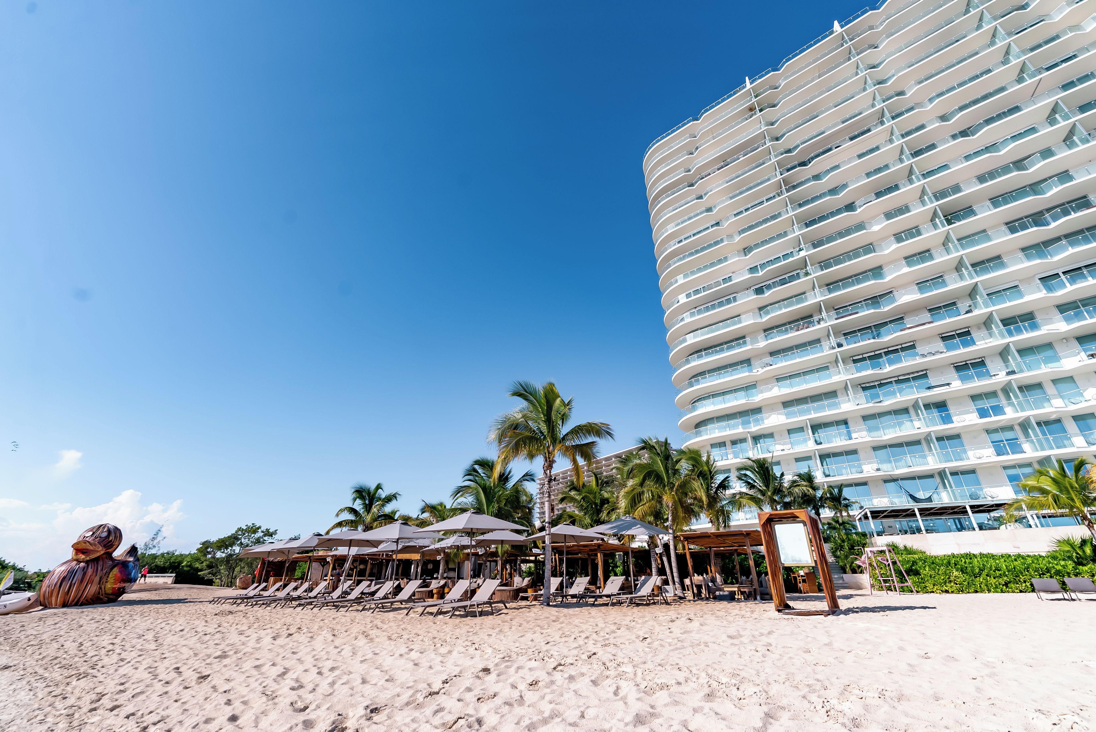 A beachfront hotel with a curved facade, multiple balconies, and a beach area with sun loungers and umbrellas.