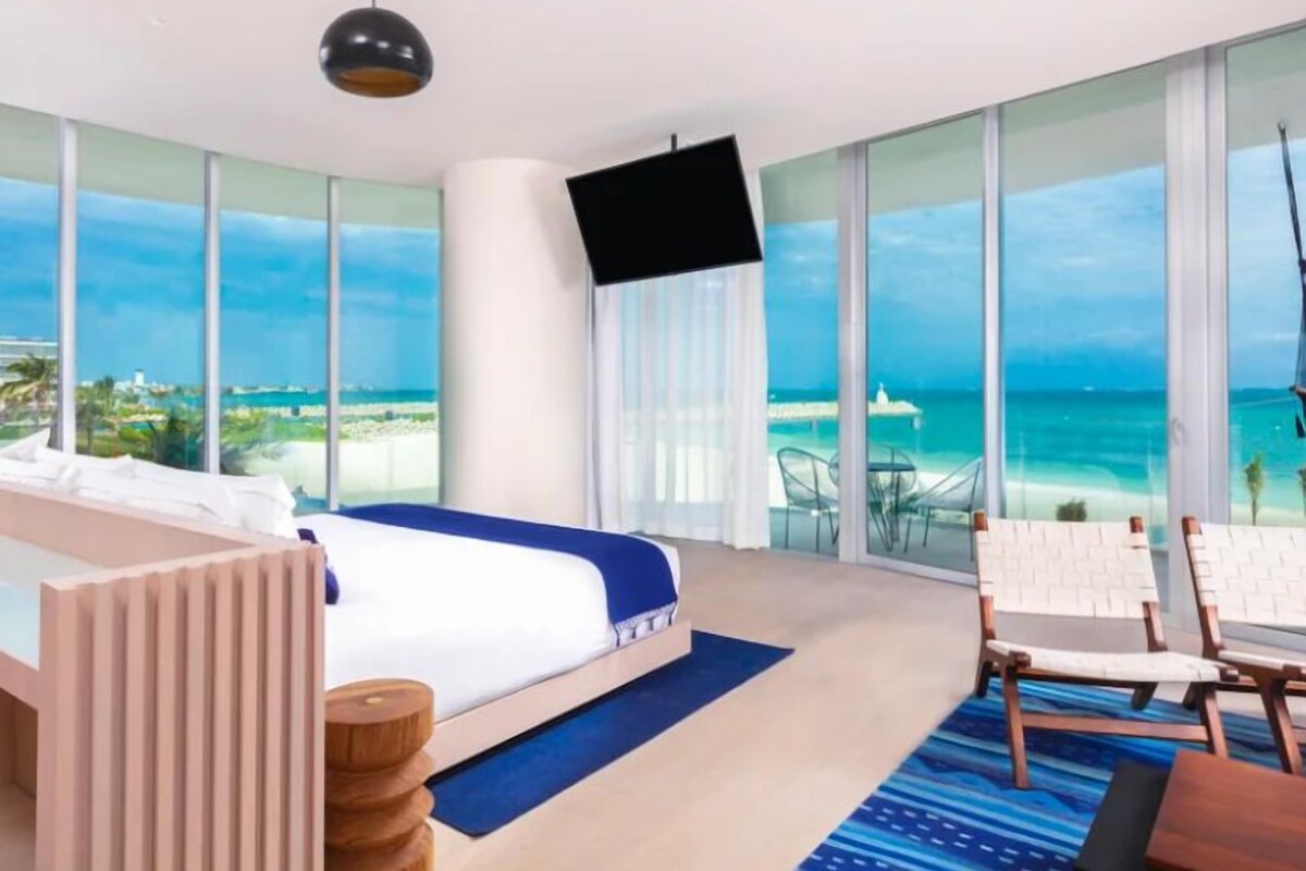 A hotel room with a large bed, a TV, a view of the ocean, and a balcony with chairs.