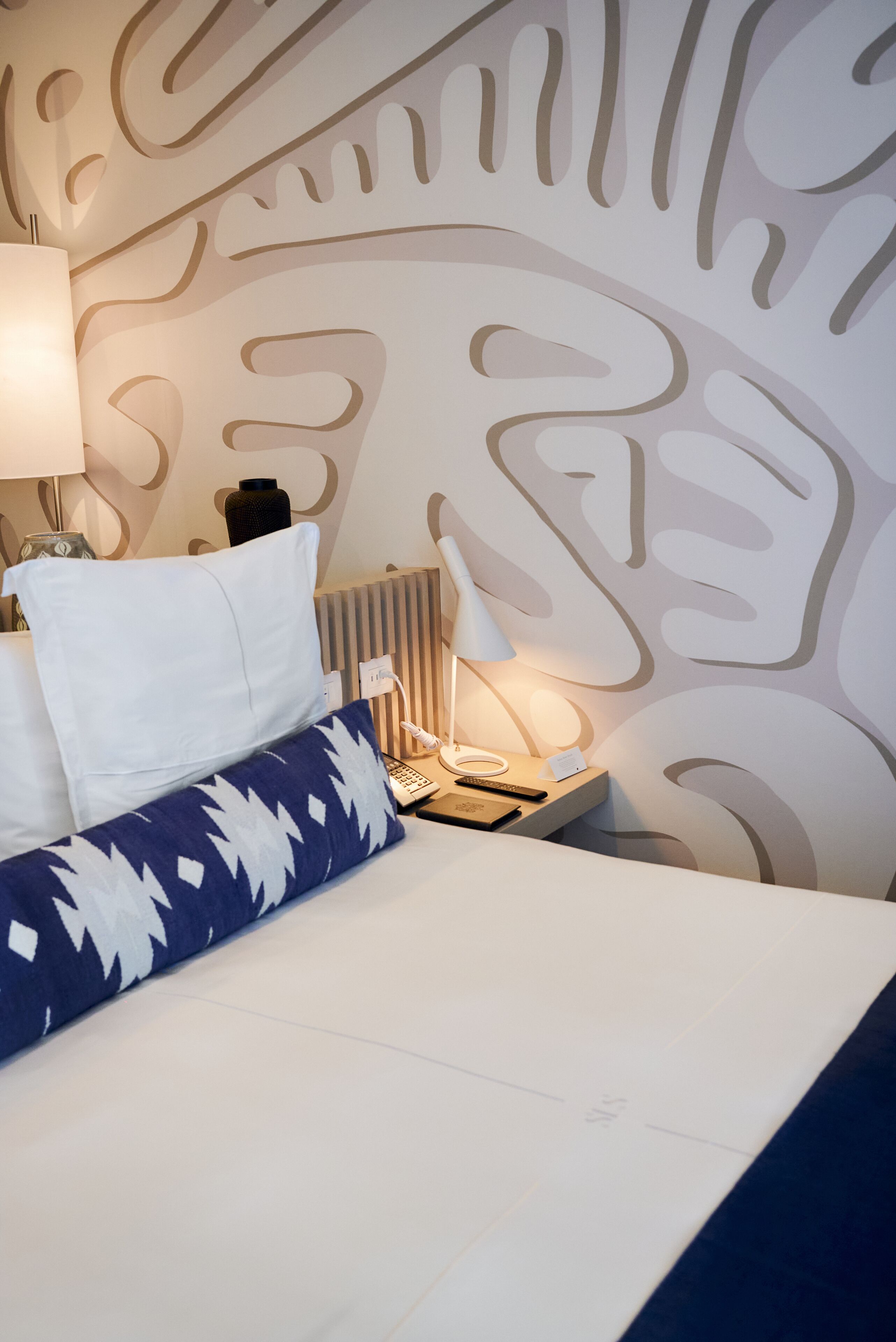 A neatly made bed with a patterned pillow, a bedside lamp, and a wall with abstract leaf designs.