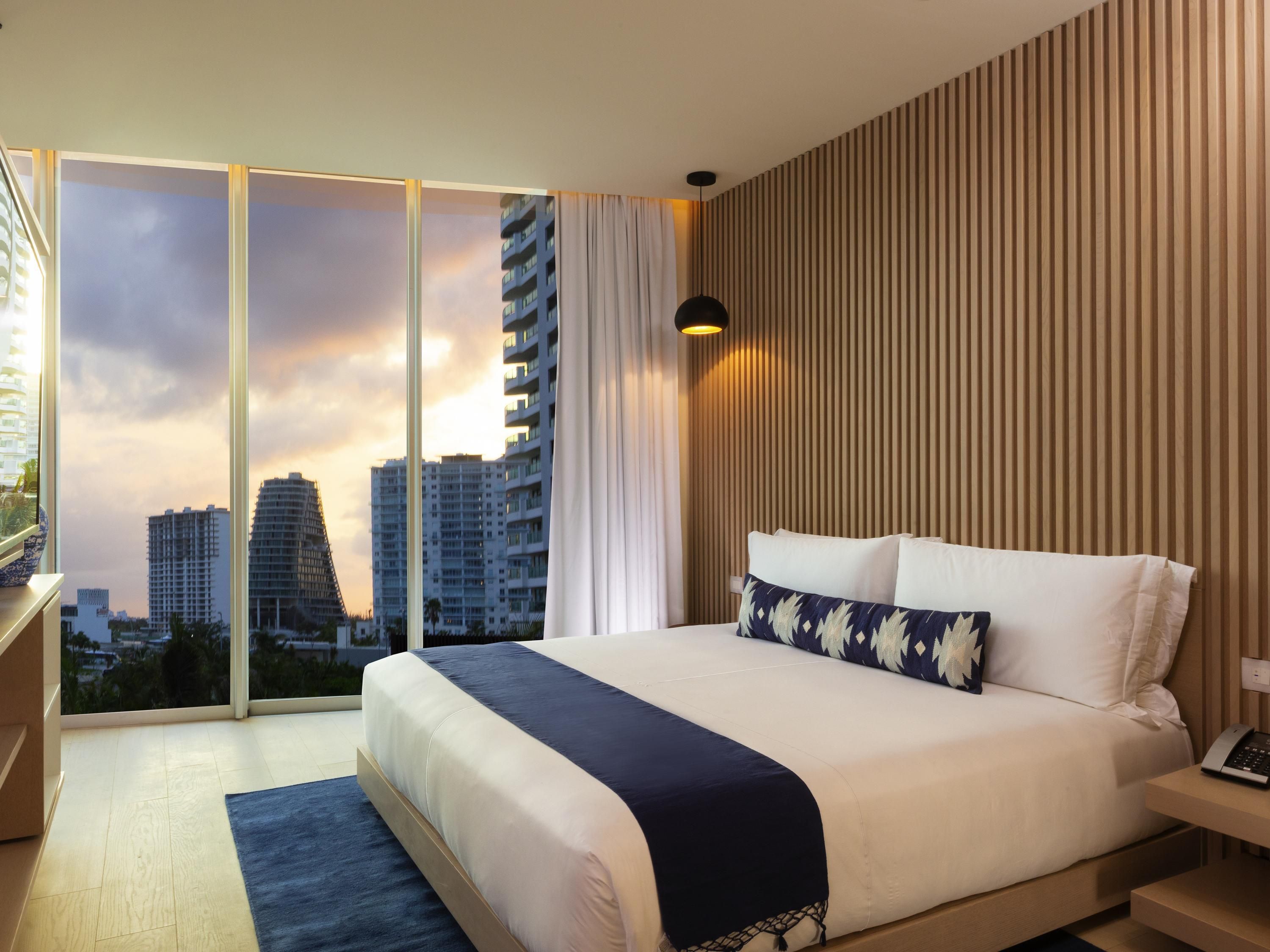 A modern hotel room with a large bed, a desk, and a view of city buildings through floor-to-ceiling windows.