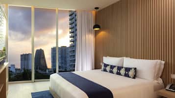 A modern hotel room with a large bed, a desk, and a view of city buildings through floor-to-ceiling windows.