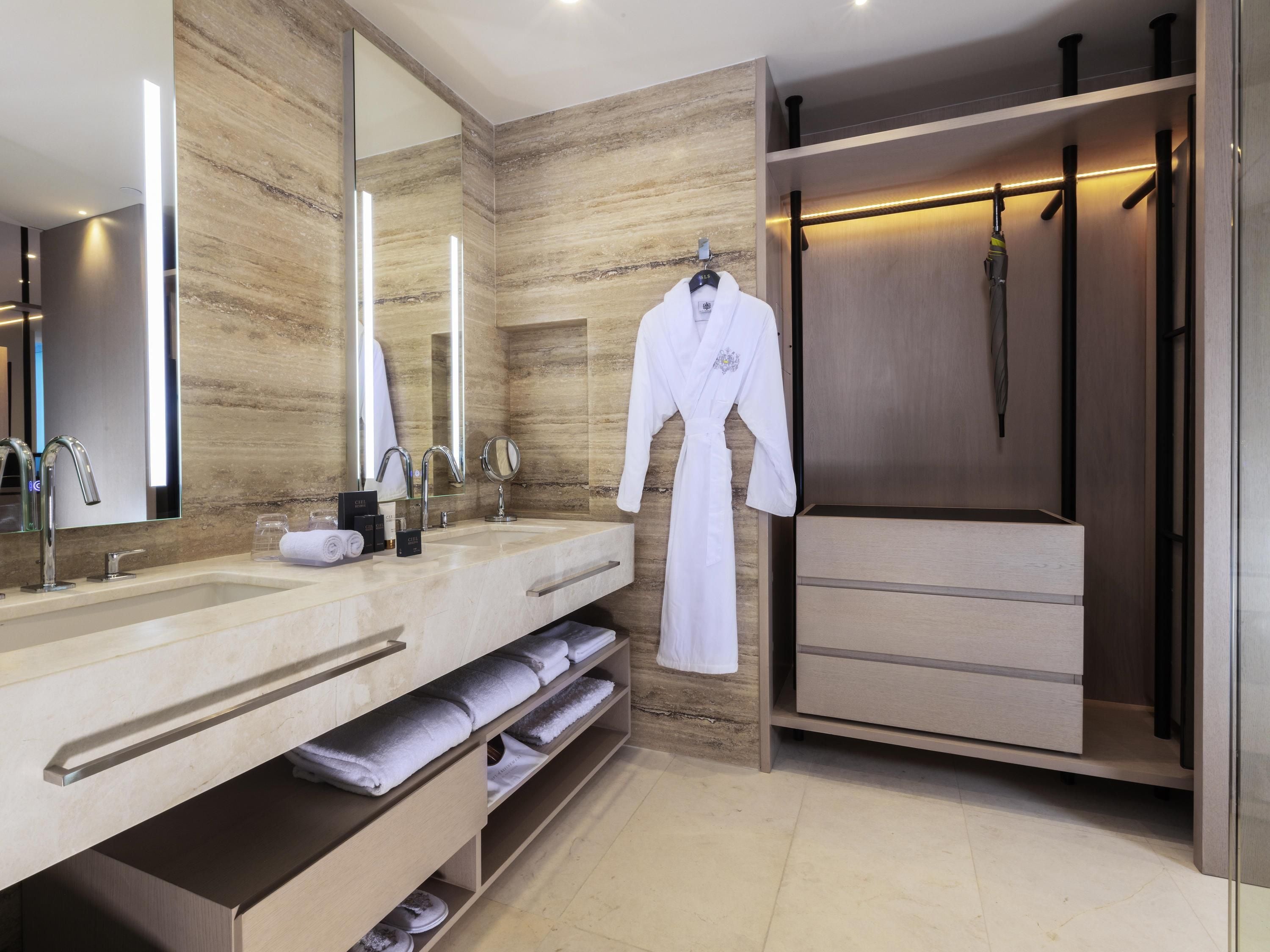Un baño moderno con tocador de mármol, un espejo grande, una bata colgada en la pared y un vestidor con detalles de madera.