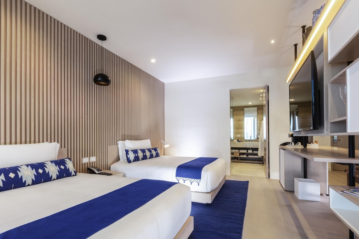 Una habitación de hotel moderna con dos camas, televisión, escritorio y baño visible a través de una puerta abierta.