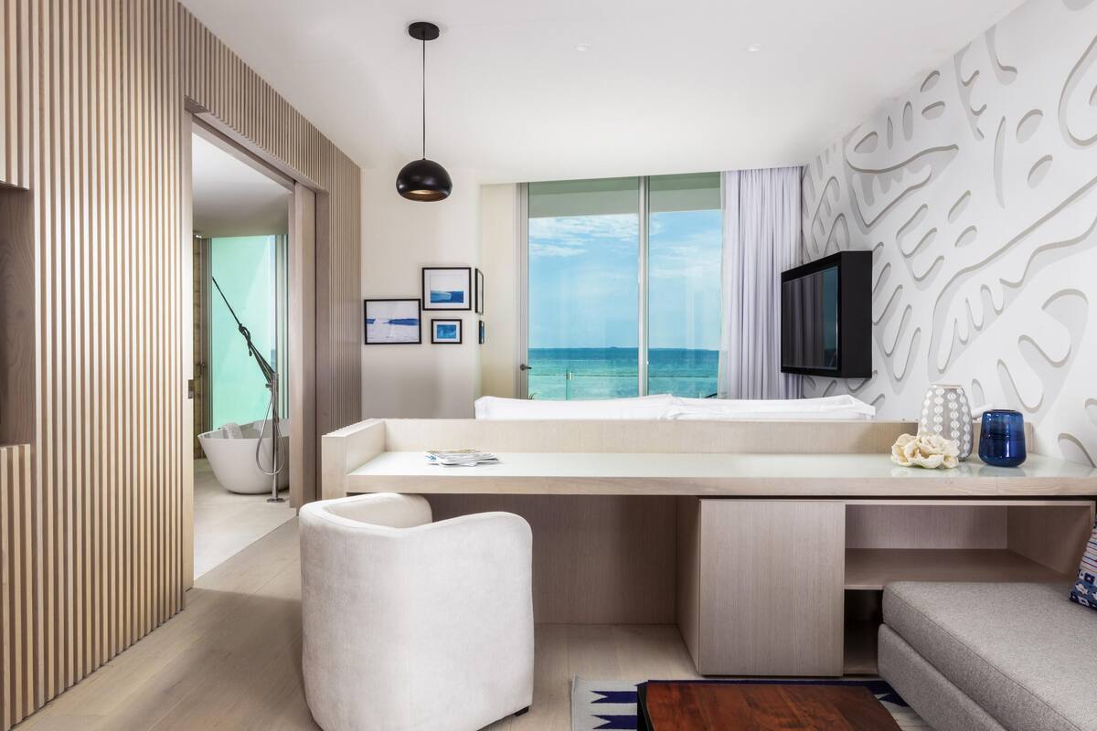 Ein modernes Hotelzimmer mit großem Fenster und Meerblick, einem flachbildfernseher an der Wand sowie einem minimalistischen Design mit einer Holzvertäfelung und einem weißen Sofa.