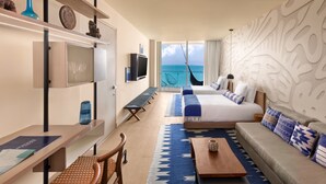 A modern hotel room with a large bed, a sofa, a desk, and a TV. There is a view of the ocean through the window.
