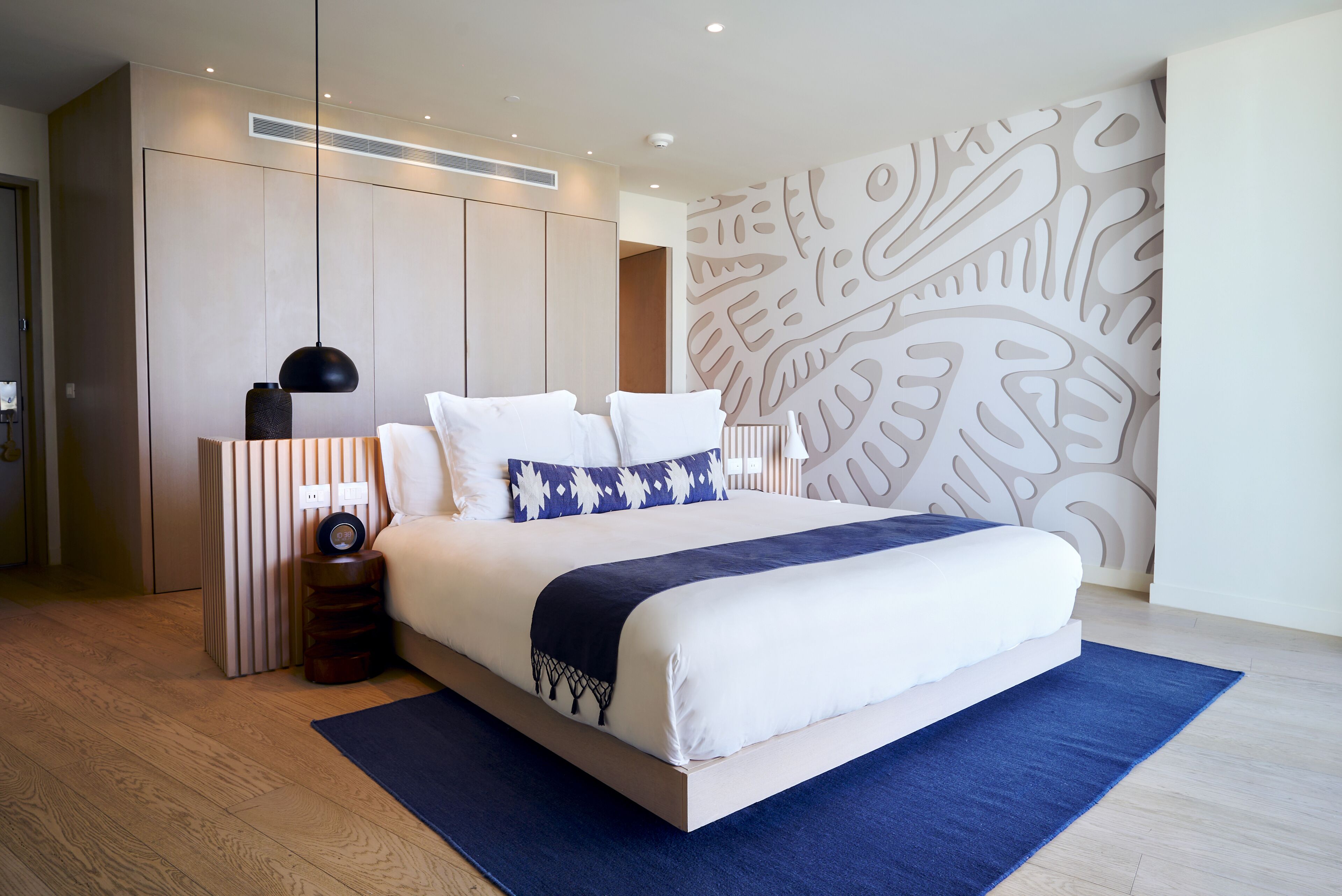 A modern bedroom with a large bed, a bedside table, a wall-mounted heater, and a decorative wall panel.
