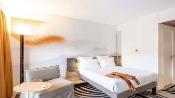 Quarto de hotel com uma cama, um pequeno sofá, mesa de cabeceira, luminária e TV fixada na parede.