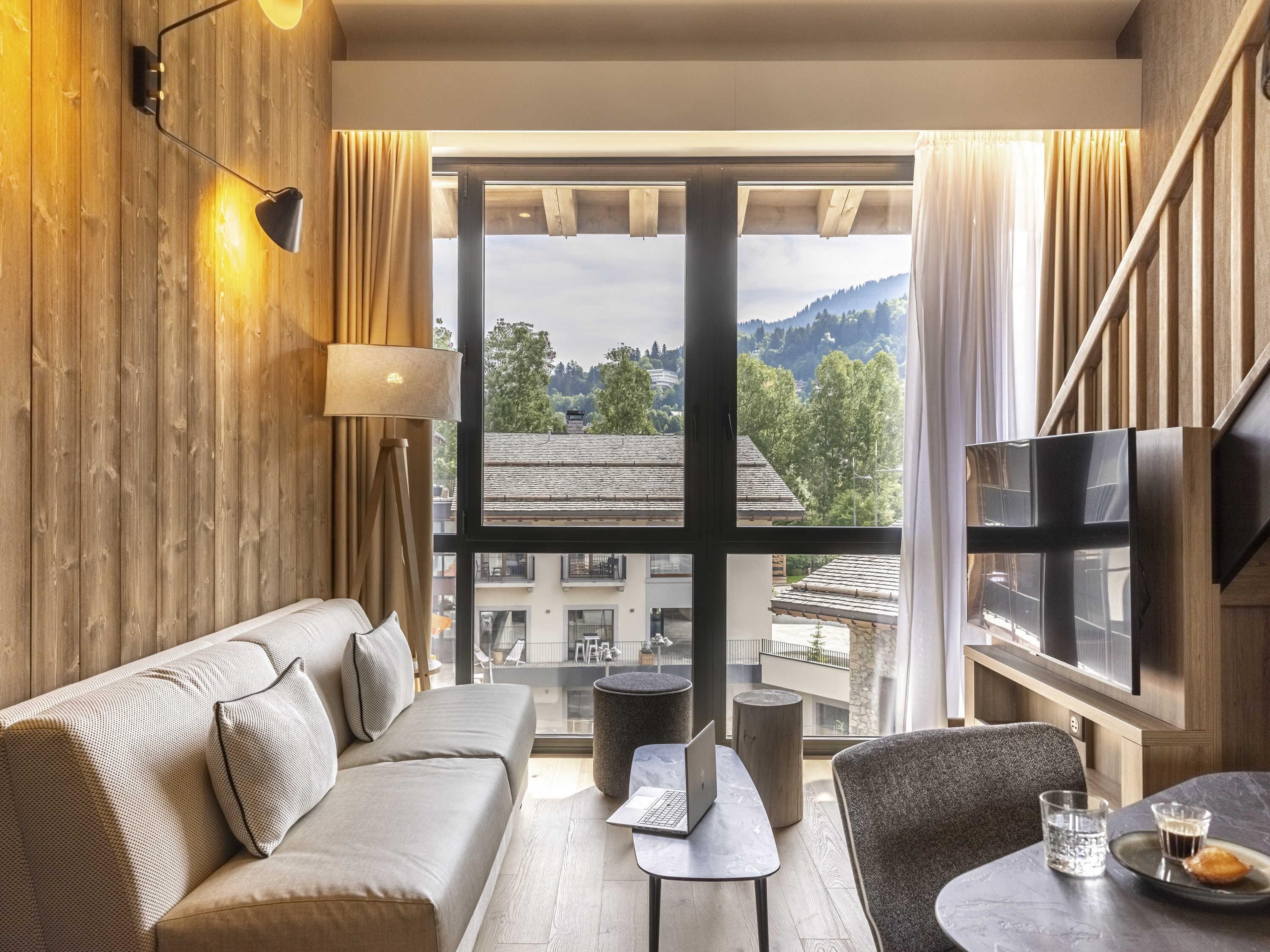 Een moderne hotelkamer met een bank, een klein tafeltje met een laptop, een stoel, een televisie en een groot raam met uitzicht op de stad.