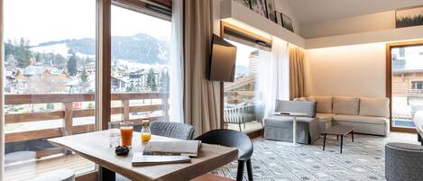 Habitación de hotel moderna con zona de comedor, sofá, televisión y vistas a las montañas.