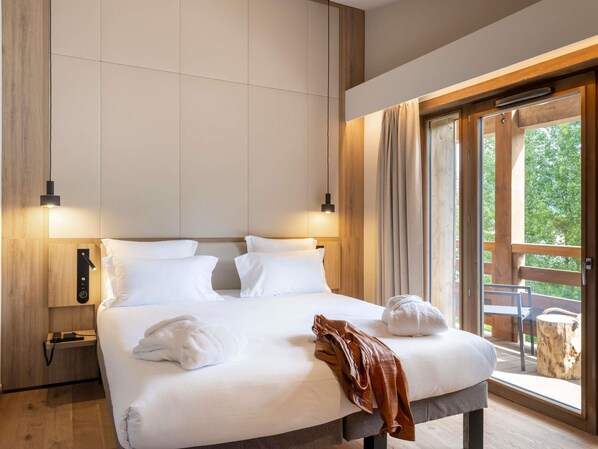 A hotel room with a bed, two bedside tables, a nightstand, a window with curtains, and a view of the outdoors. - Novotel Megève Mont-Blanc (Megeve)