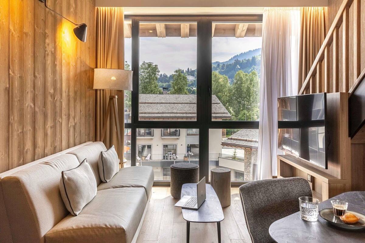 A modern hotel room with a sofa, a small table, a TV, and a large window offering a city view.