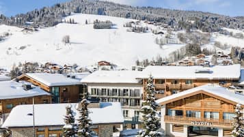 A snow-covered hotel with multiple wooden buildings, a ski slope in the background, and a clear sky.